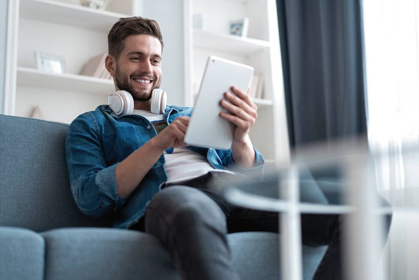 Attractive young man is using a tablet and smiling while lying on sofa at home. - Фото, изображение