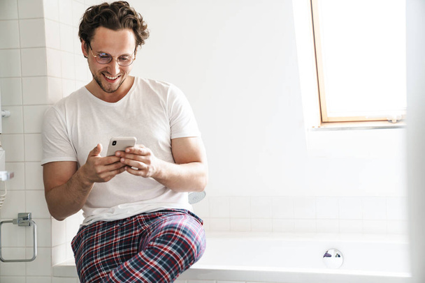 Smiling young man wearing pajamas sitting on a bathtub, using mobile phone - Valokuva, kuva