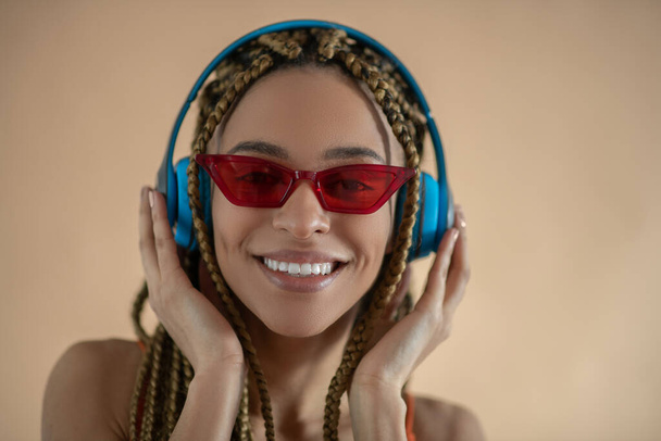 Smiling young African American female in red eyeglasses, touching blue headphones and listening to music - Fotografie, Obrázek