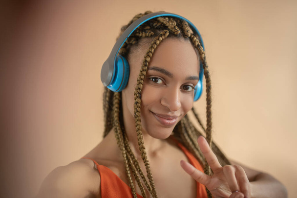 Smiling young African American female in blue headphones, showing peace sign, making selfie - Fotó, kép