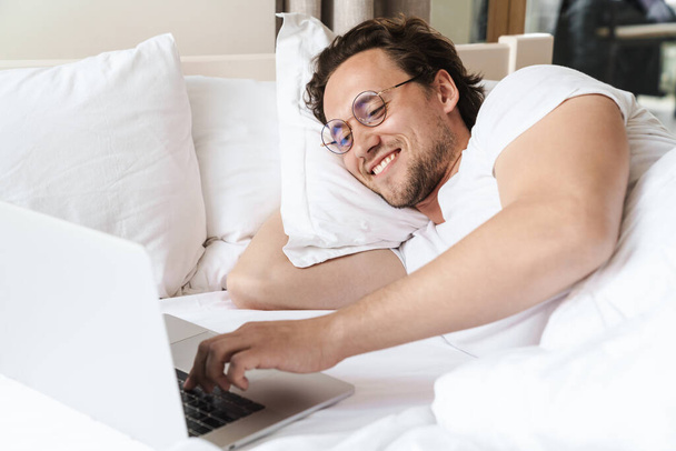 Smiling young man laying on a pillow in bed, using laptop computer - Foto, Imagem