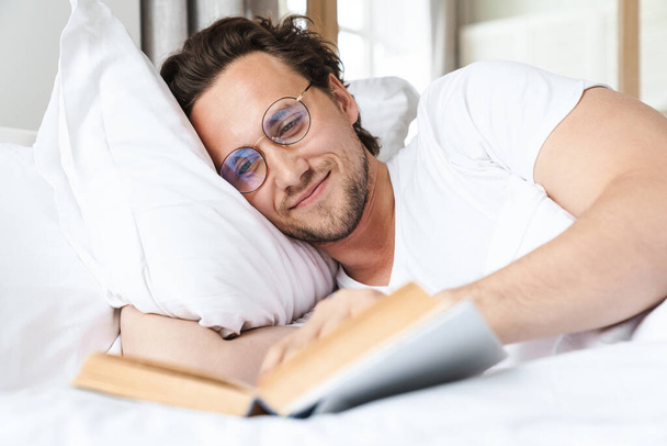 Smiling young man laying on a pillow in bed, reading a book - Fotoğraf, Görsel