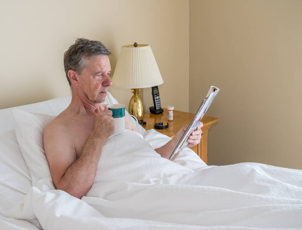 Egyedülálló fehér idős férfi ferde ágyban és magazinból olvas - Fotó, kép