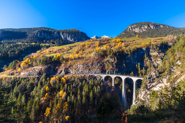 Ohromující pohled na Landvasser Viadukt v odpoledním slunci za podzimního dne s alpami a barevnými stromy, Kanton Grisons, Švýcarsko - Fotografie, Obrázek