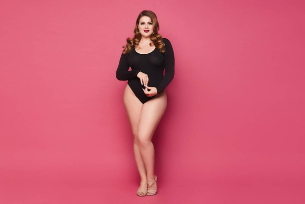 Young plump woman with makeup and trendy hairstyle wearing a black bodysuit and posing at the pink background, isolated. - Zdjęcie, obraz