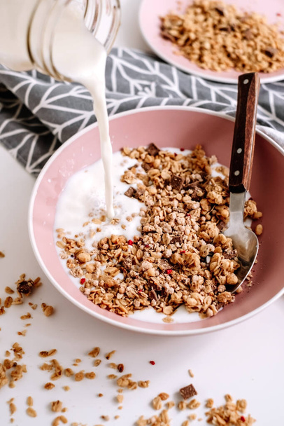 Granola muesli with fresh milk and pieces of chocolate in a pink plate on a white table. Healthly food, breakfast concept. High quality photo - Zdjęcie, obraz