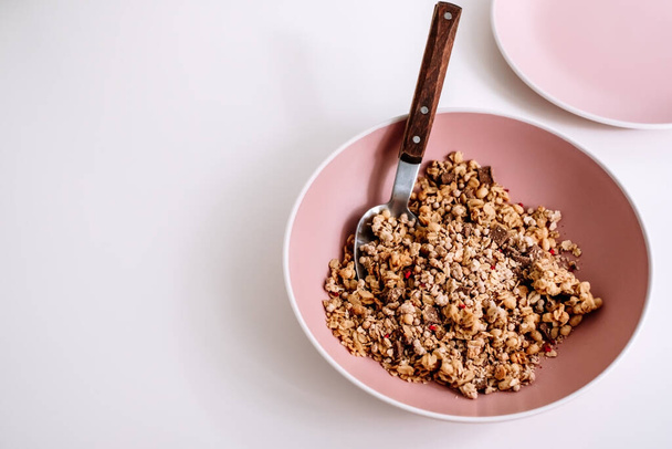 Granola muesli with pieces of chocolate in a pink plate on a white table. Healthly food, breakfast concept. High quality photo - Zdjęcie, obraz