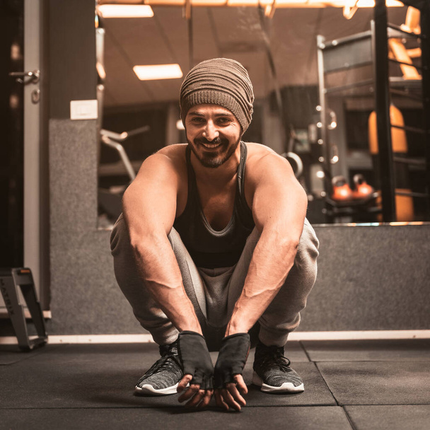 Χαμογελώντας αθλητικός άνθρωπος με μάλλινο καπέλο κάθεται στο γυμναστήριο μετά την προπόνηση. Κοίτα κατευθείαν στην κάμερα.. - Φωτογραφία, εικόνα
