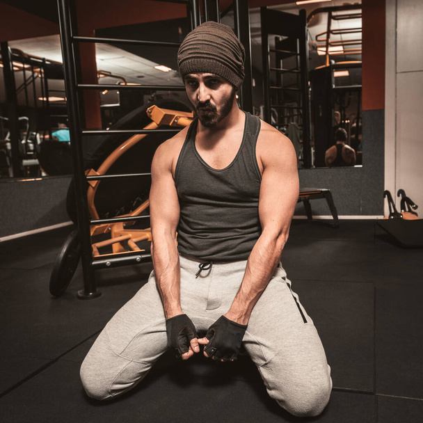 Muscular tired bodybuilder guy showing his muscular body taking a break from running or exercise sitting in the gym. Fitness young sporty man resting in workout. Train in a healthy way. - Photo, Image