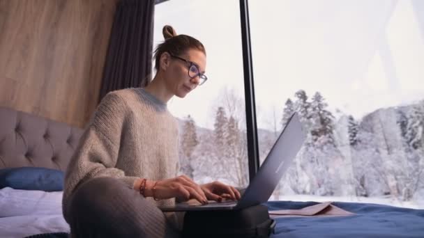 A woman freelancer with glasses sits on a bed in a country house with panoramic windows behind which there is a snowy forest. Girl with laptop computer does remotely work while isolated - Záběry, video