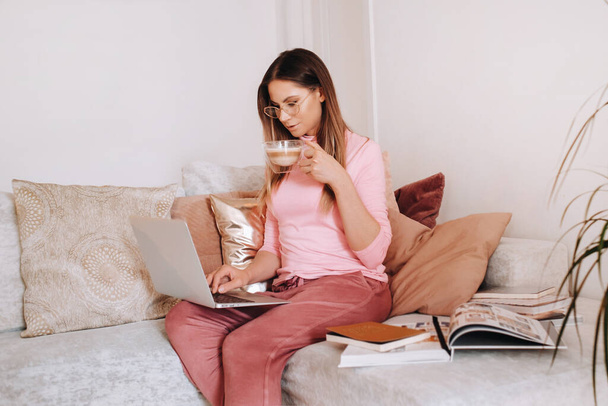a girl in the morning in pajamas at home working on a laptop with drinking coffee, a girl self-isolated at home and resting on the couch and watching a laptop.Household chores. - Foto, Imagen