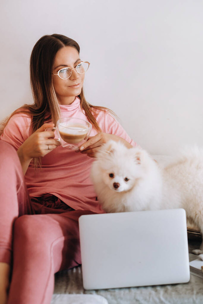 a girl in pajamas at home is working on a laptop with her dog Spitzer, the dog and its owner are resting on the couch and watching the laptop.Household chores. - Photo, Image