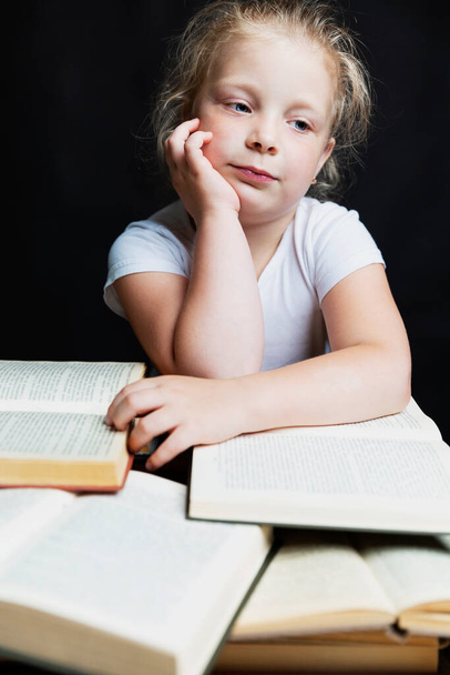 Грустная маленькая девочка сидит с грудой книг. Знания и образование. Чёрный фон. Вертикаль
. - Фото, изображение