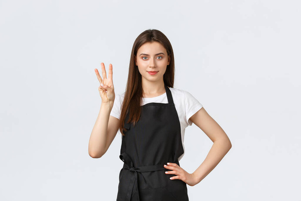Grocery store employees, small business and coffee shops concept. Cheerful young female barista in black apron showing number three, smiling determined, working in cafe - Photo, Image