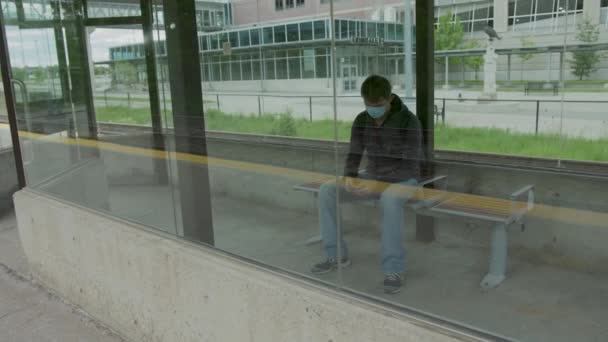 High quality 4k video of a young man who is waiting for a transportation in a glass bus stop or station and holding his cell phone in his hands. Man is texting on a station while waititng for a train or bus and wearing blue medical face mask  - Πλάνα, βίντεο
