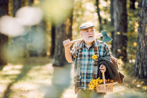 Bearded man relax in forest. Smiling old forester with axe and flowers. Happy man with beard and mustache hold axe. Elderly male walk in forest. Bearded man in hat on a background of trees. - Foto, Imagem
