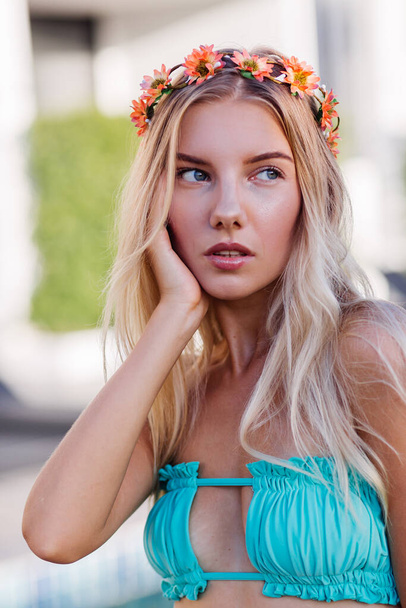 Portrait of young happy blonde long hair woman in yellow trendy sunglasses, blue sexy bikini and flower wreath on head, summer mood, stylish fashion look 2020.  Female on vacation by swimming pool. - Foto, Imagen