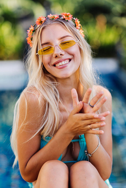 Portrait of young happy blonde long hair woman in yellow trendy sunglasses, blue sexy bikini and flower wreath on head, summer mood, stylish fashion look 2020.  Female on vacation by swimming pool. - Φωτογραφία, εικόνα