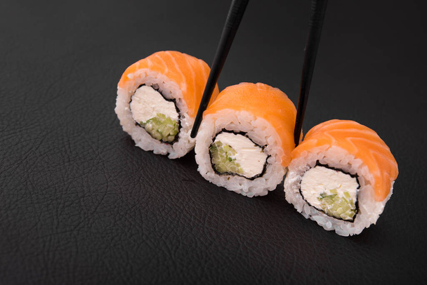 close-up foto van een sushi roll met zalm en komkommer wordt gehouden met eetstokjes op een zwarte achtergrond - Foto, afbeelding