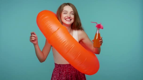Joyful, extremely happy girl in dress dancing inside big rubber ring and drinking refreshing juice, laughing carefree, relaxing on party with cocktail. indoor studio shot isolated on blue background - Video