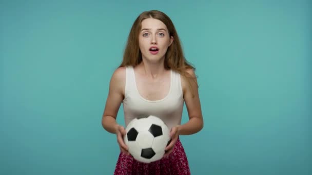 Pretty girl football fan holding soccer ball in her hands and eagerly watching sports game, in anticipation of winning goal, shouting for joy, victory. indoor studio shot isolated on blue background - Video