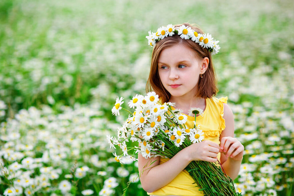 Ένα μικρό κορίτσι στη φύση με ένα στεφάνι μαργαρίτες από λουλούδια στο κεφάλι της και ένα μπουκέτο στα χέρια της. Πεδίο των χαμομηλιών σε μια ηλιόλουστη μέρα του καλοκαιριού. - Φωτογραφία, εικόνα
