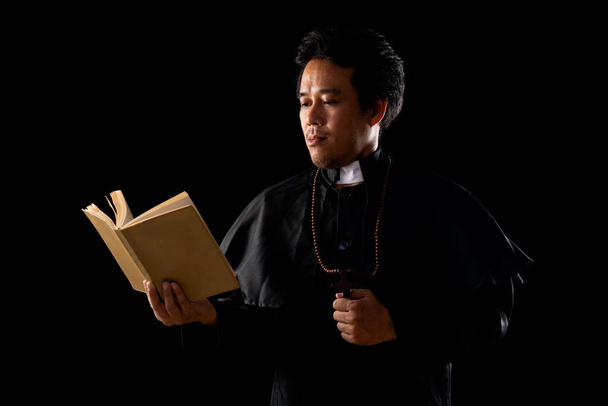 Sacerdote jovem com uniforme preto segurando Bíblia velha em fundo preto
 - Foto, Imagem