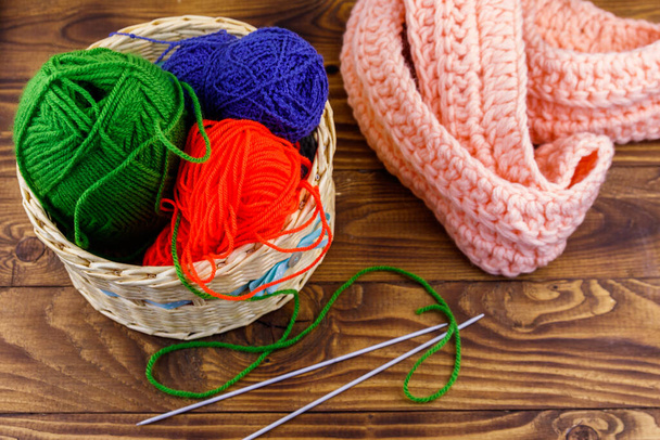 Плетеная корзина с красочными вязальными нитями, вязальными спицами и вязаным шарфом на деревянном столе - Фото, изображение
