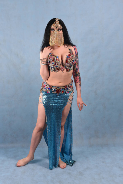 A beautiful dancer in an Oriental dress with a veil and a red skirt with full Breasts on a blue background in the Studio - Photo, image