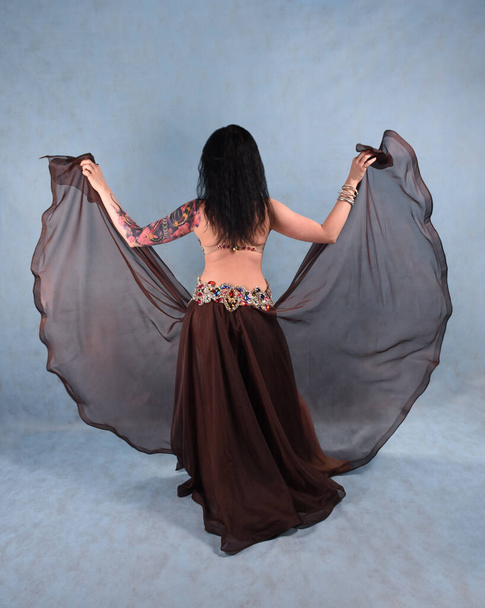 A beautiful dancer in an Oriental dress is dancing in the Studio on a blue background waving a brown skirt and dress - Photo, Image