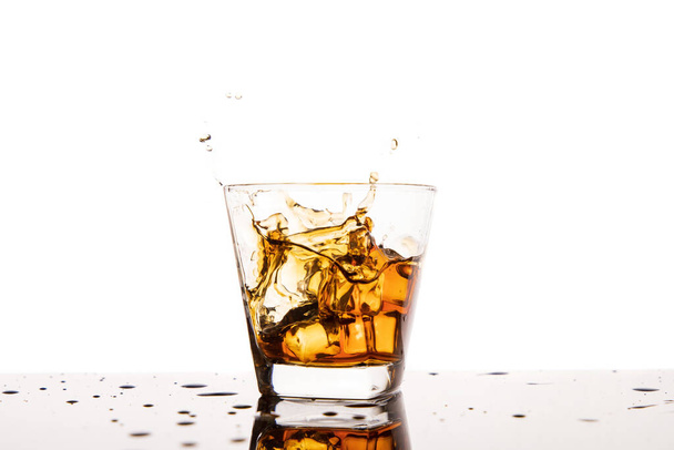 アイススプラッシュ付きのウィスキーグラス。白に隔離された氷でウイスキーを振りかけるガラス - 写真・画像
