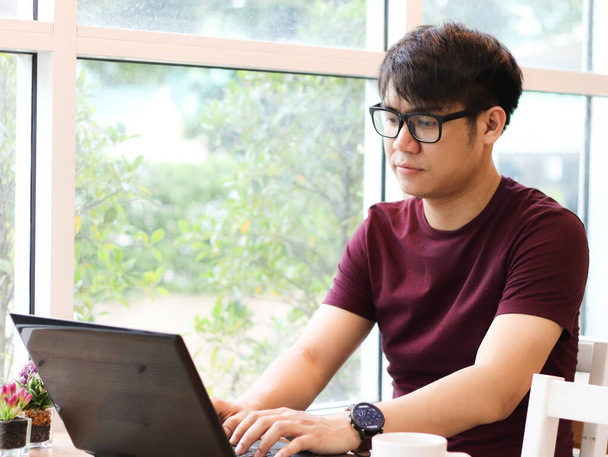 Asian young man wearing eye glasses sitting in the coffee shop looking  thoughtfully at screen of  computer laptop on the table , - Photo, Image