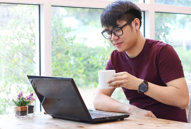 Nuori aasialainen mies silmälasit istuu ikkunan ääressä vihreä puu ulkona tilalla kuppi kahvia lukeminen postia tietokoneen kannettava näyttö ja hymy.Nuori mies työskentelee kahvilassa tietokoneen kannettava tietokone pöydällä. - Valokuva, kuva