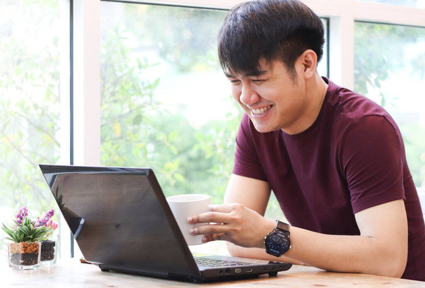 Asian young man sitting by the window in coffee shop with computer laptop on the table , looking at computer screen and laughing. - Foto, afbeelding