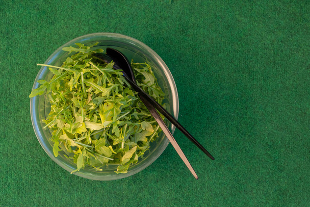 Schüssel mit frischem gesunden Rucola oder Rucola für einen Salat mit Servierutensilien von oben auf grün mit Copyspace betrachtet - Foto, Bild