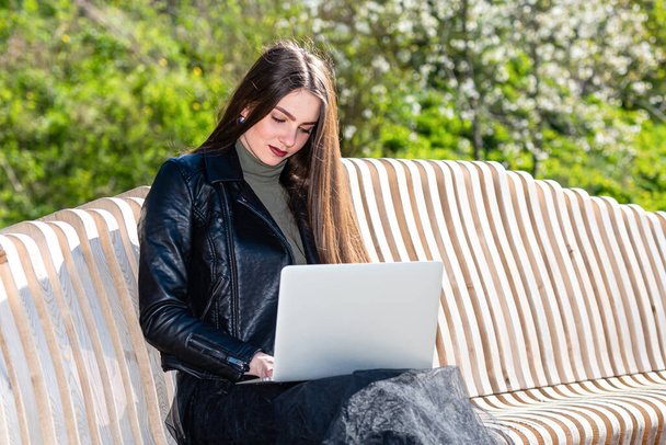 νεαρή κομψή γυναίκα κάθεται στον πάγκο στο πάρκο και εργάζονται σε φορητό υπολογιστή σε μια ηλιόλουστη και θυελλώδη μέρα, η έννοια της εργασίας έξω από το γραφείο - Φωτογραφία, εικόνα