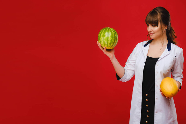 a female doctor nurse in a white coat with fruit in her hands poses on a red background, melon, watermelon - Fotoğraf, Görsel