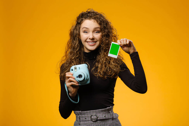 Šťastná dívka s okamžitou filmovou kamerou izolované na žlutém pozadí. Model drží prázdný rámec s maketovým prostorem na obrázku - Fotografie, Obrázek