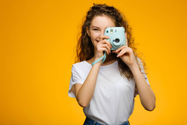 Pretty girl with instant camera isolated on yellow background. Model wearing casual white shirt and has long curly hair. - Zdjęcie, obraz