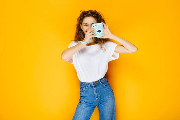 Pretty girl with instant camera isolated on yellow background. Model wearing casual white shirt and has long curly hair. - Foto, afbeelding