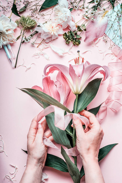 Female hands making lovely pink lily flowers bouquet on pastel  table with florist decoration equipment, top view. Creative Florist workspace and flowers arrangement.  Festive holiday concept. - Fotó, kép