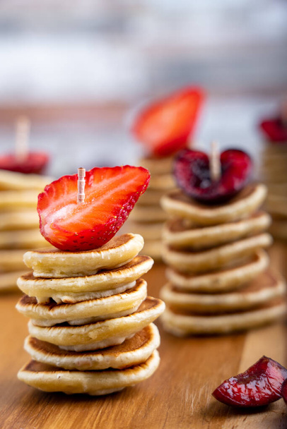 Stacks of mini pancakes with topping made of strawberry and cherry slices standing on wooden cutting board at kitchen - Фото, изображение