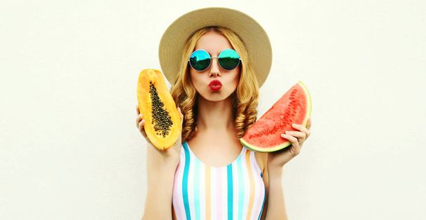 close up portrait of attractive young woman with fruits slice of watermelon and papaya blowing red lips sending sweet air kiss wearing straw hat, sunglasses on white background - 写真・画像