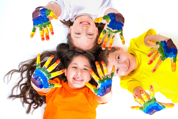 Troje szczęśliwych i uśmiechniętych dzieci patrzy w kamerę i wyciąga ręce w farbach. Tęcza na dłoniach. Koncepcja szczęśliwego dzieciństwa i dnia dla dzieci - Zdjęcie, obraz