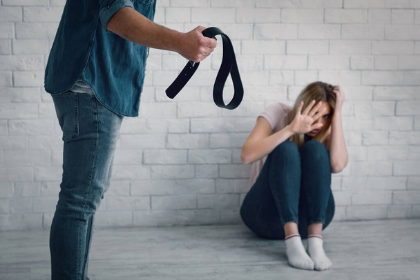 Victim and domestic violence. Aggressive man waving belt at wife sitting on floor - Фото, изображение