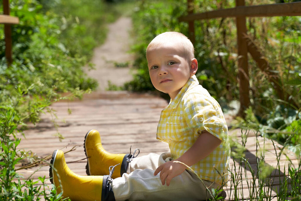 a funny little boy in yellow rubber boots sits on the bridge. - Foto, imagen