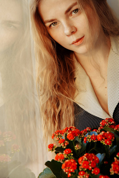 A beautiful girl is sitting by window. She hasKalanchoe flowers on her hands.  - Photo, image