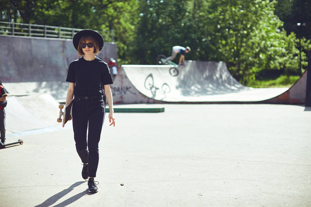Pretty funky teenage girl carrying longboard walking in skatepark in sunglasses, hat and black clothes, for ad or blog - Foto, Imagem