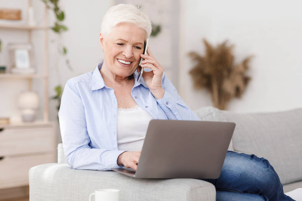 Moderní seniorka pracující na notebooku a mluvící doma na mobilu - Fotografie, Obrázek