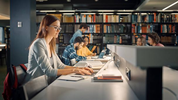 University Library: Talented Caucasian Girl Sitting at the Desk, Uses Laptop, Writes Notes for the Paper, Essay, Study for Class Assignment. Diverse Group of Students Learning, Studying for Exams. - Foto, imagen
